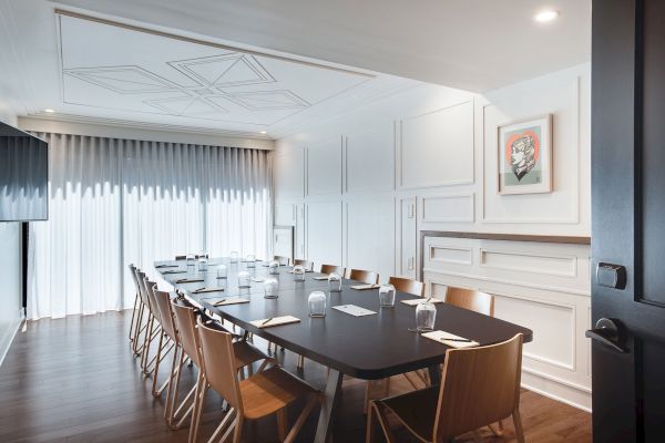 A modern conference room with a large table, wooden chairs, and a piece of artwork on the wall, lit by natural light from the windows.