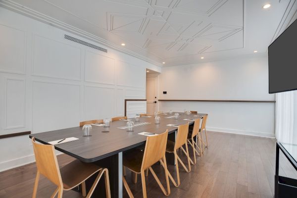 A modern conference room with a long table, wooden chairs, glass cups, and notepads, featuring a large TV screen on the wall.