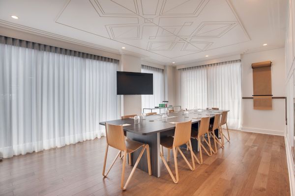 A modern conference room with a long table, chairs, wall-mounted TV, and large windows with curtains, featuring a geometric ceiling design.