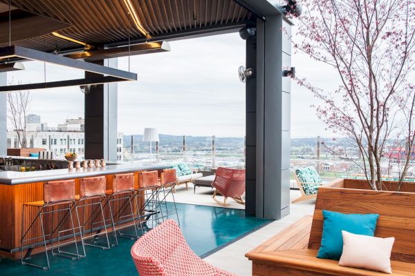 Rooftop lounge with a bar, comfy seating, and city views. Modern decor includes bar stools and cushioned benches under a partially open roof.