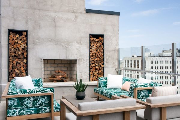 A rooftop patio with green-patterned seating, fireplace, stacked firewood, and potted plant, against a city backdrop under a clear sky.