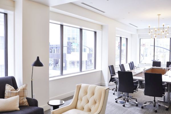 The image shows a modern office space with large windows, a conference table, swivel chairs, and seating areas with couches and a floor lamp.