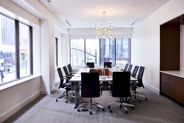 A modern conference room with a long table, black chairs, large windows, and a chandelier, creating a professional setting.