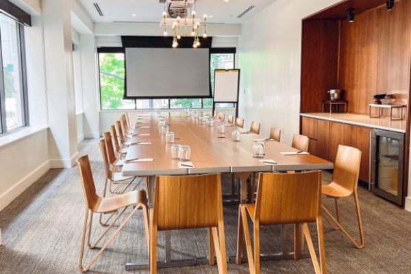 A conference room with a long table, wooden chairs, a projector screen, and a kitchenette. Bright windows and modern decor complete the space.