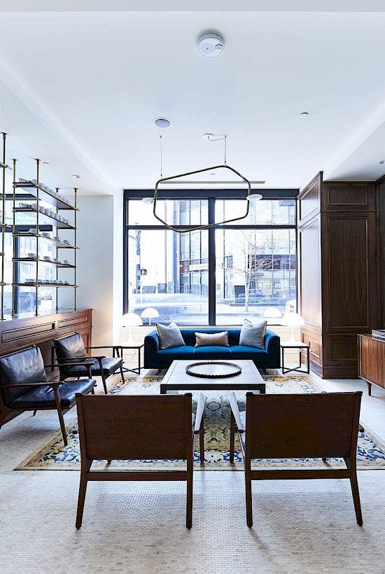 A modern living room with a blue sofa, chairs, and shelves, featuring large windows and wooden accents.