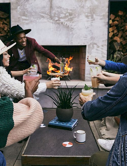 A group of people sit around a coffee table near a fireplace, holding drinks and wearing hats, creating a cozy and social atmosphere.