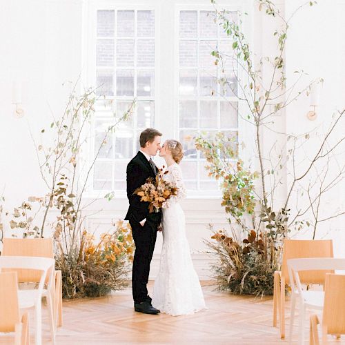 A couple stands closely in a bright, airy room decorated with plants and flowers, in front of a large window.