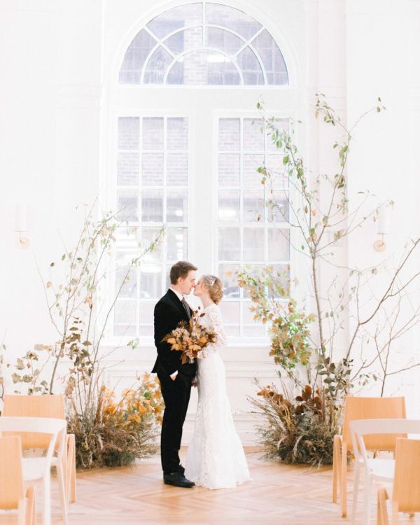 A couple stands closely in a bright, airy room decorated with plants and flowers, in front of a large window.