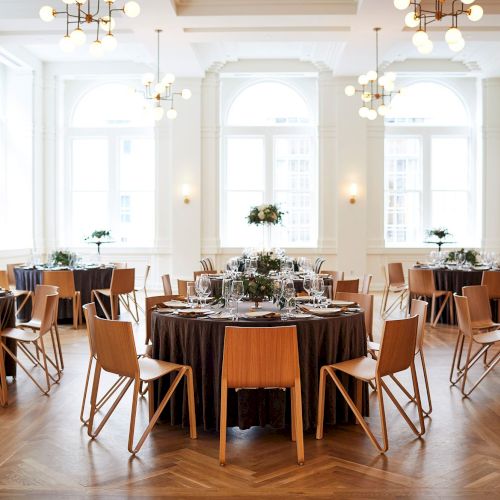 The image shows an elegant dining room with round tables set for an event, featuring wooden chairs and large windows in a bright space.