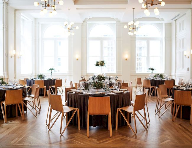 The image shows an elegant dining room with round tables set for an event, featuring wooden chairs and large windows in a bright space.