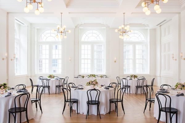 A bright, elegant dining room with round tables, white tablecloths, black chairs, and floral centerpieces, lit by chandeliers.