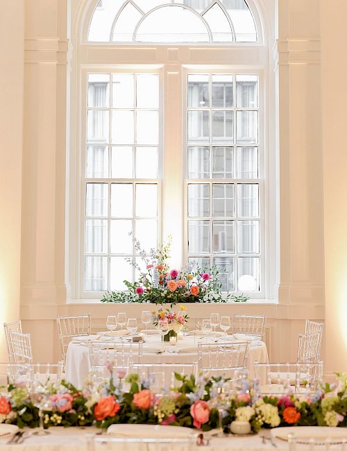 An elegantly set table with floral arrangements in a bright room, featuring a large arched window in the background.