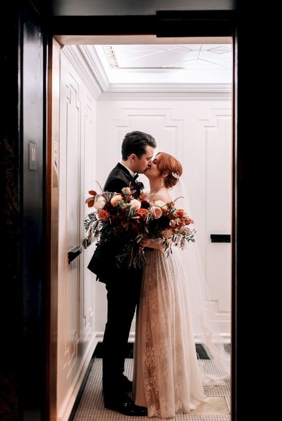 A couple is sharing a kiss in an elevator, holding a bouquet of flowers, dressed in formal attire.