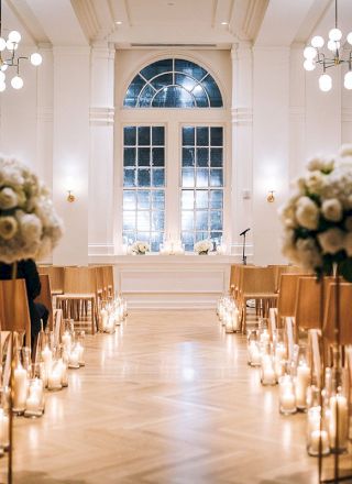 An elegant indoor wedding setup features floral arrangements, rows of wooden chairs, and soft candle lighting in a beautifully lit room.