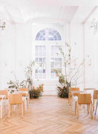 A bright room with wooden chairs, plants, and a large window featuring an arch design lets in natural light, creating an elegant atmosphere.