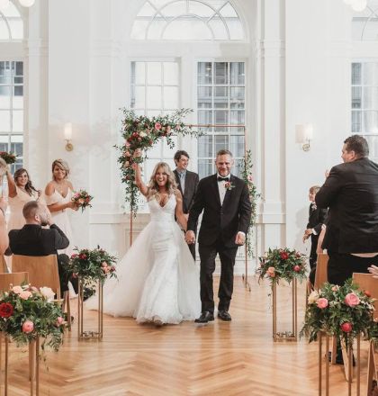 A bride and groom walk down the aisle to applause in a bright, elegant venue with floral decorations and seated guests.