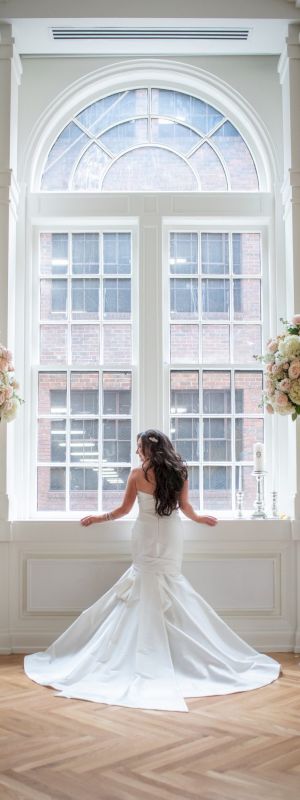 A bride in a wedding dress stands before a large window with tall floral arrangements on each side, in a bright, elegant setting.