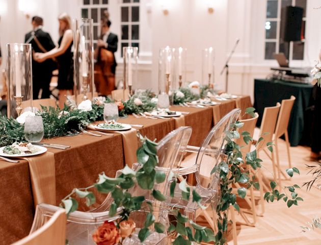 Elegant dining setup with tables decorated with greenery, candles, and a few people in the background in a stylish room.