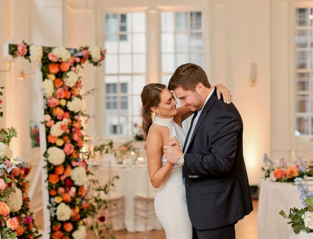 A couple is sharing a dance in a beautifully decorated room with floral arrangements and elegantly set tables.