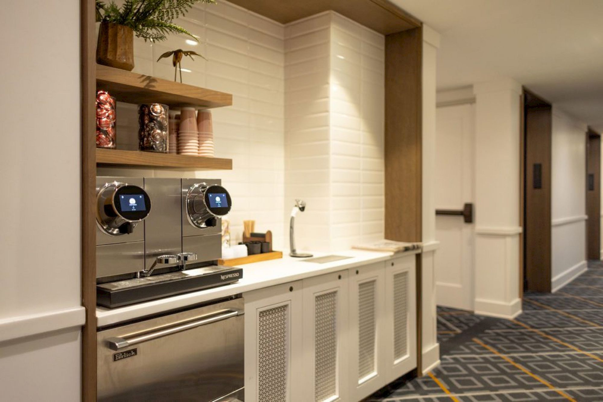 The image shows a coffee station in a hallway with shelves, coffee cups, and a modern coffee machine.