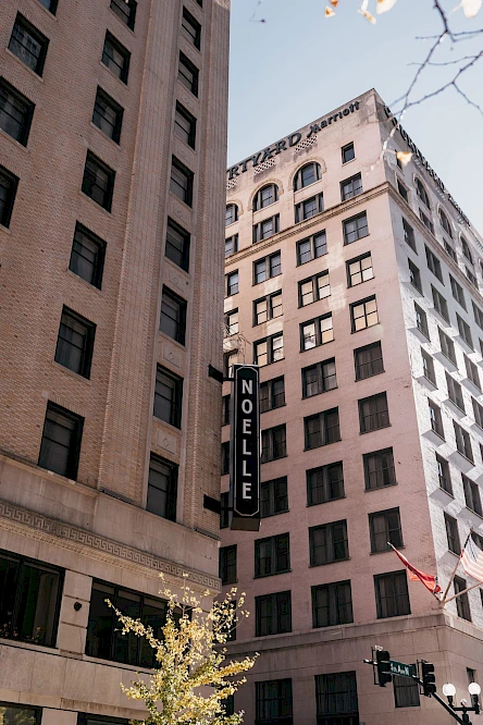 The image shows two tall buildings, with one displaying a "NOELLE" sign. There's a tree in the foreground and a red flag on the right.