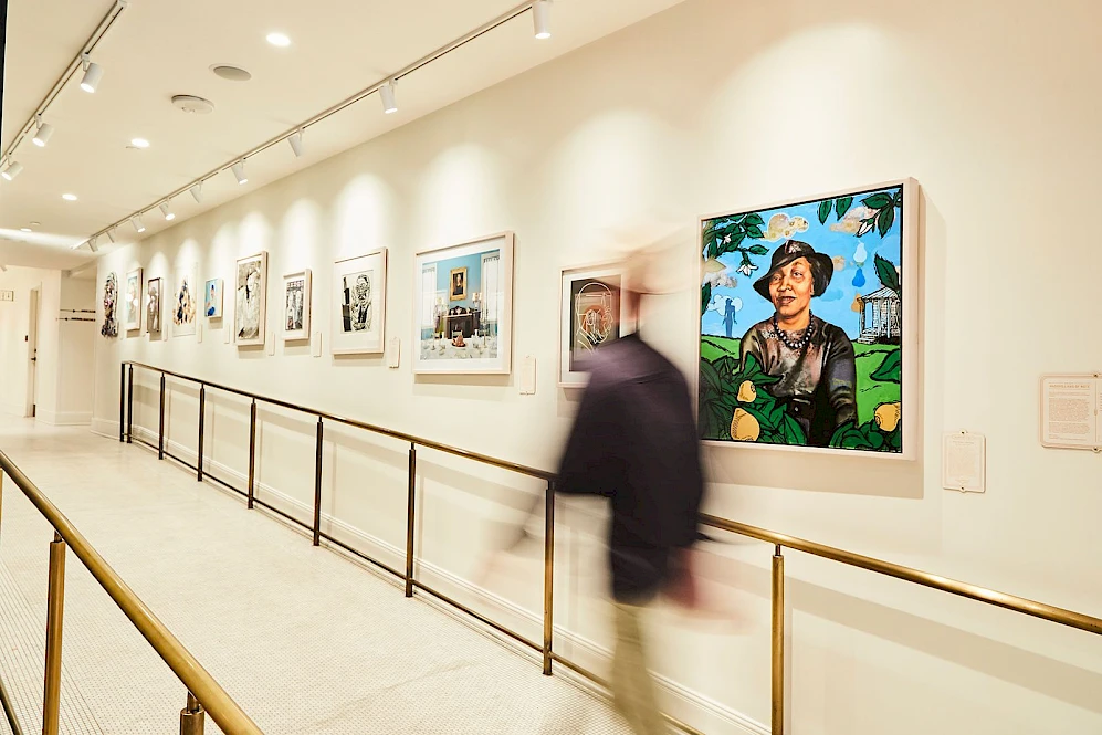 A person walks through an art gallery, with paintings on display along a well-lit corridor featuring a central railing.