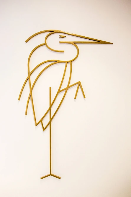 A minimalist line art depiction of a bird, resembling a heron or egret, crafted from thin metal rods mounted on a wall.