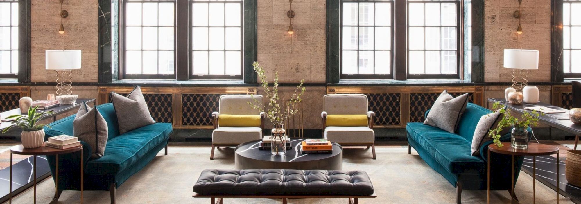 A stylish living room with teal sofas, armchairs, and a round coffee table. Large windows and lamps add light to the space.