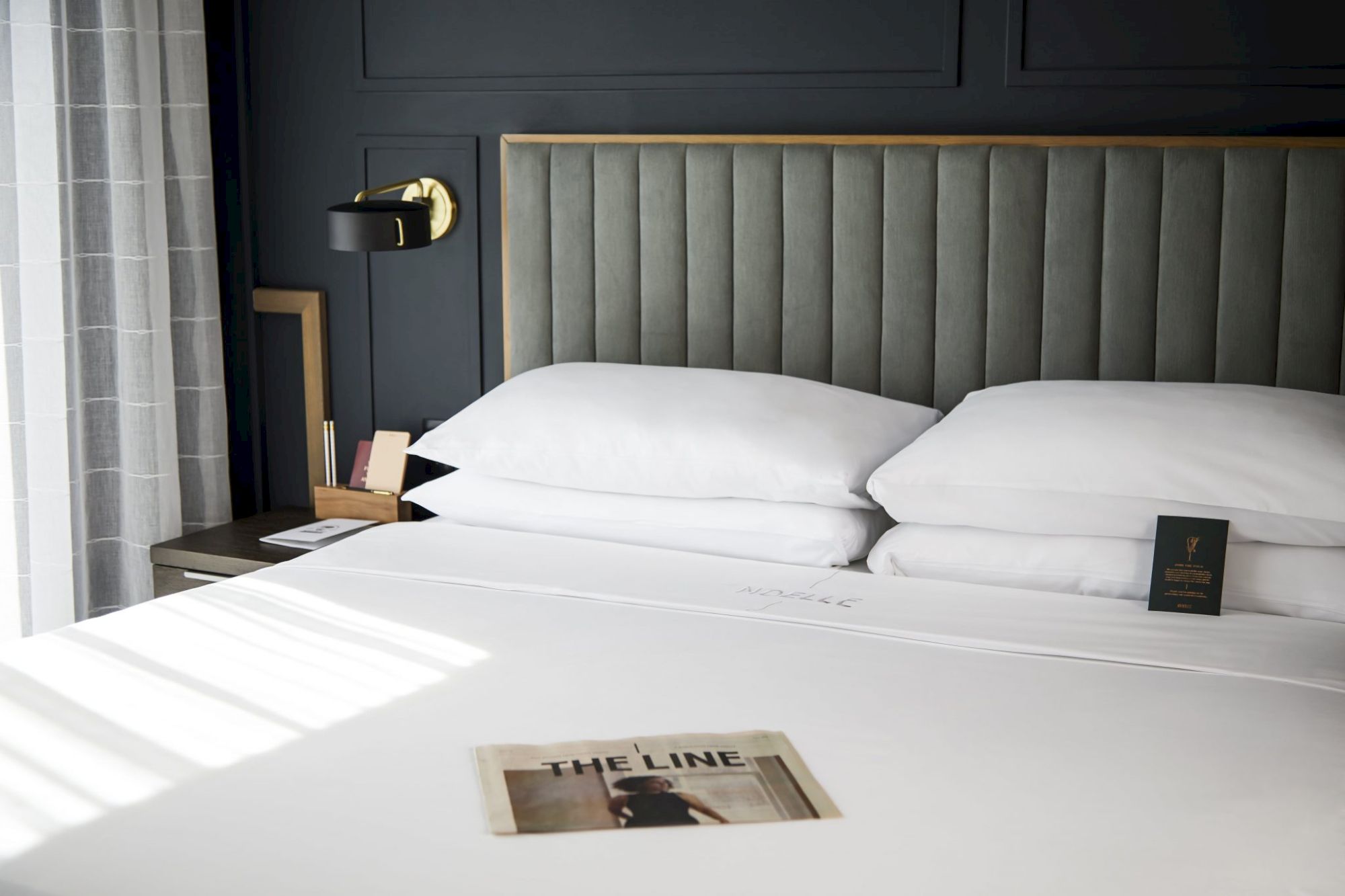 A neatly made bed with two pillows, a magazine on the duvet, a headboard, and a wall lamp beside a curtain-lined window completes the scene.