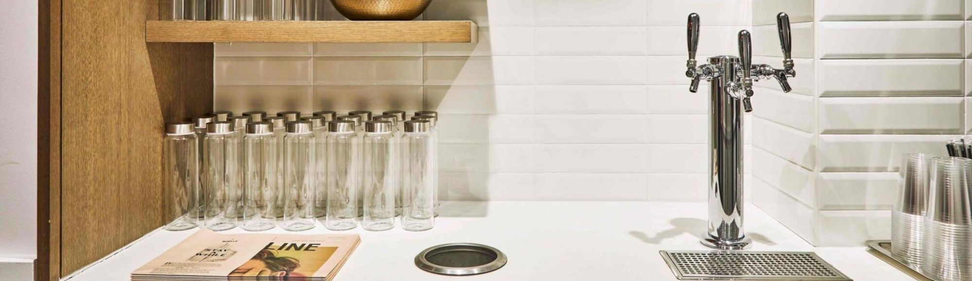 The image shows a counter with a drink dispenser, clear bottles, plastic cups, a wooden shelf, and a magazine, all against a white tiled backdrop.