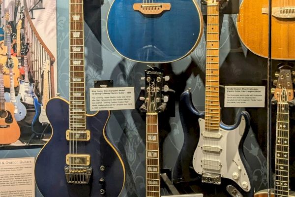 The image shows a display of various electric and acoustic guitars, including a blue and black model, in a museum or showroom setting.