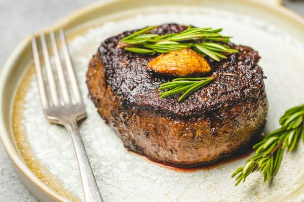 A seasoned steak garnished with herbs and a slice of garlic on a plate, accompanied by a fork.