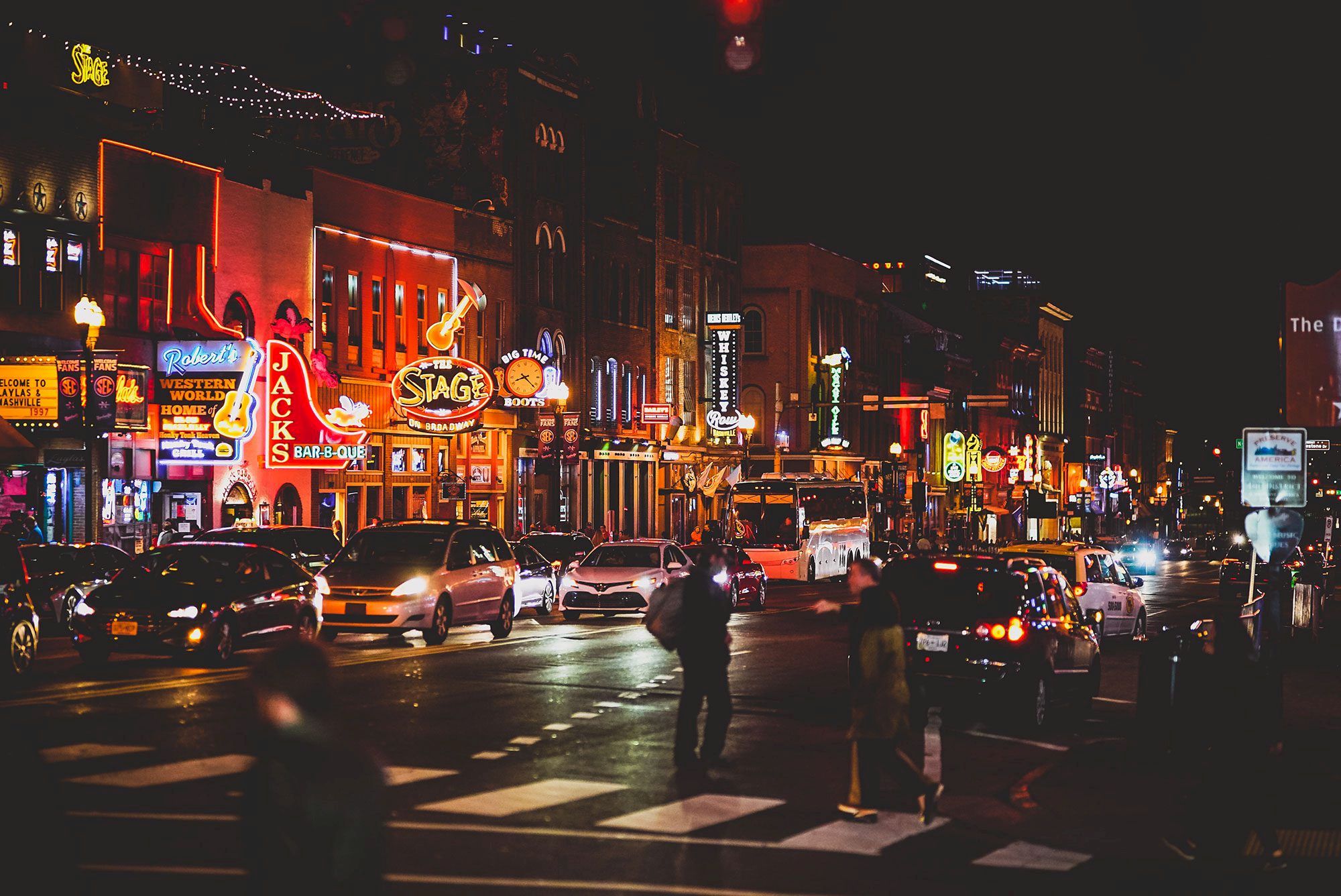 A bustling city street at night with bright neon signs, cars, and people crossing.