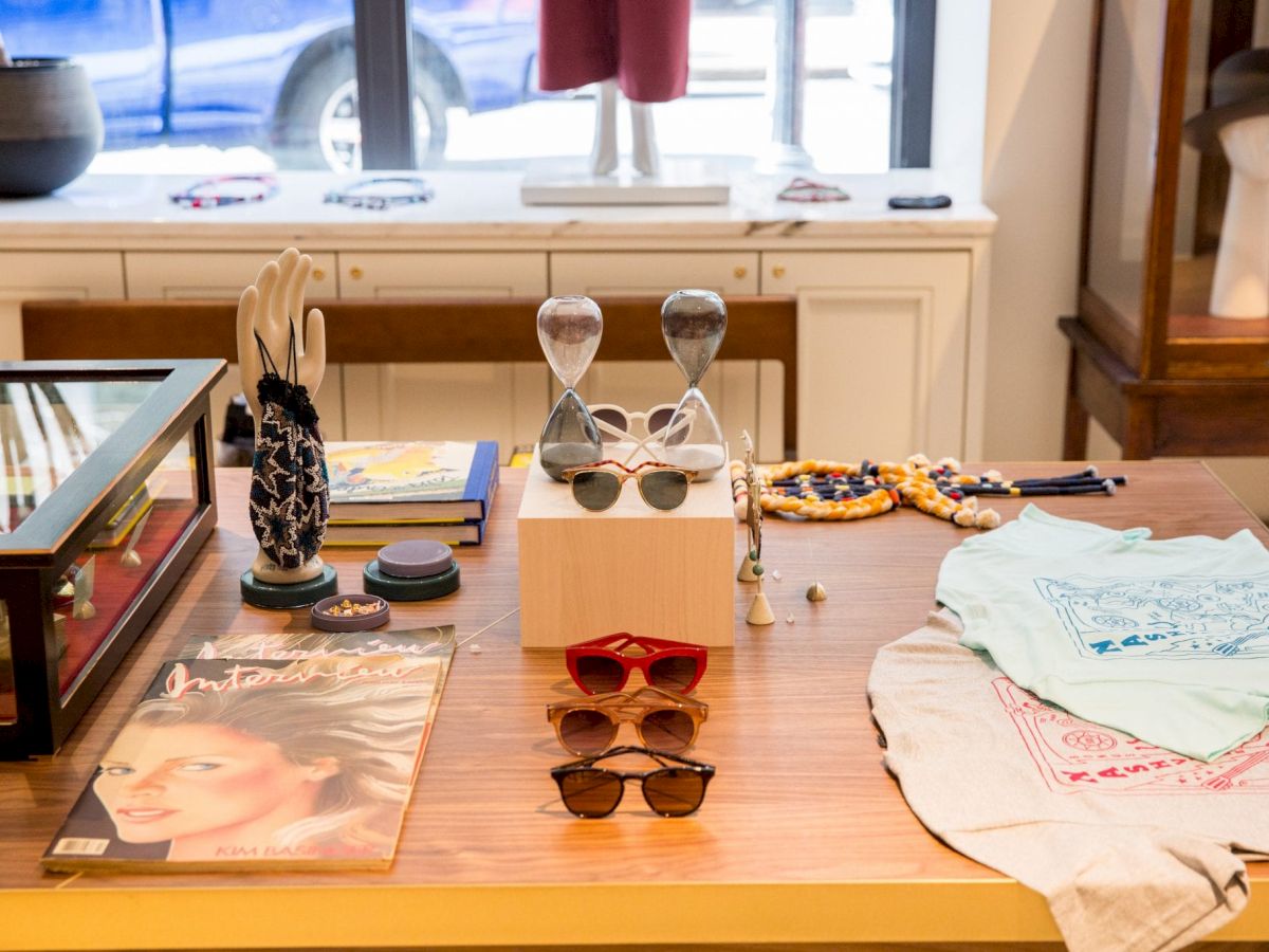 The image shows a display table with sunglasses, a magazine, decorative items, jewelry, and folded clothing in a shop setting.