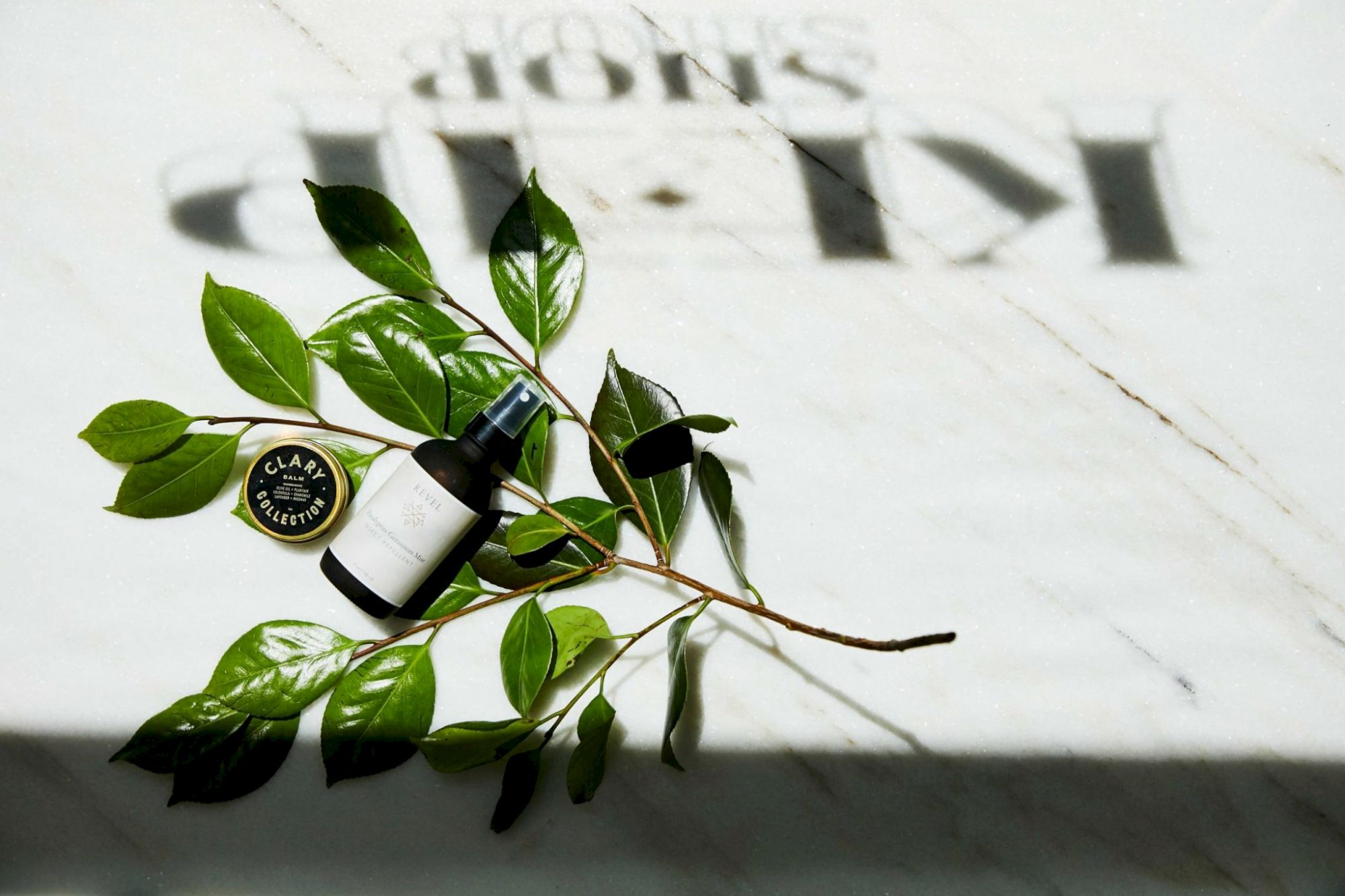 A skincare or cosmetic product bottle and a small container are placed on leaves on a marble surface with a shadowed 