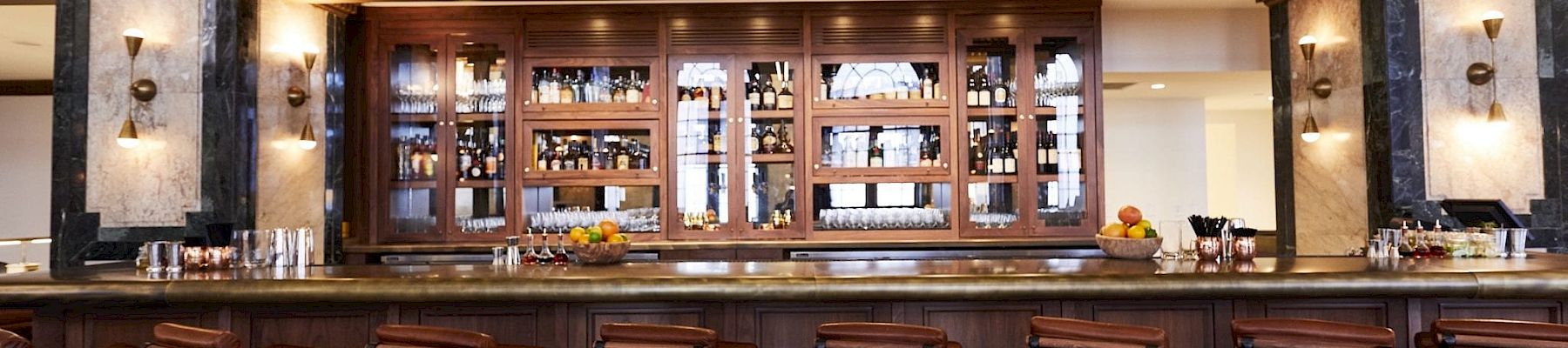 An elegant bar interior with high stools, a wooden counter, and shelves full of bottles, set in a high-ceilinged room with ornate decor.