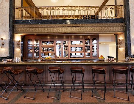 An elegant bar interior with high stools, a wooden counter, and shelves full of bottles, set in a high-ceilinged room with ornate decor.