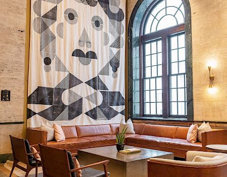 A stylish lounge with a large abstract face artwork, leather seating, chairs, a coffee table, and a tall window enhancing natural light.