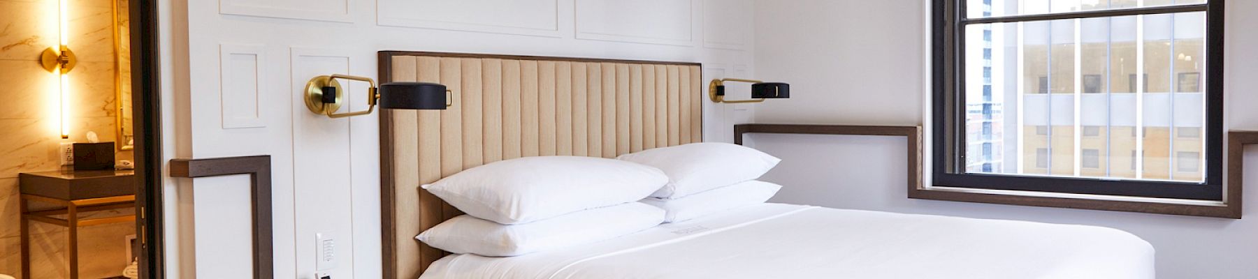 A modern hotel room with a neatly made bed, a window, and a small side table beside it. Through the door, a bathroom is partially visible.