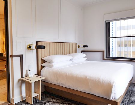 A modern hotel room with a neatly made bed, a window, and a small side table beside it. Through the door, a bathroom is partially visible.