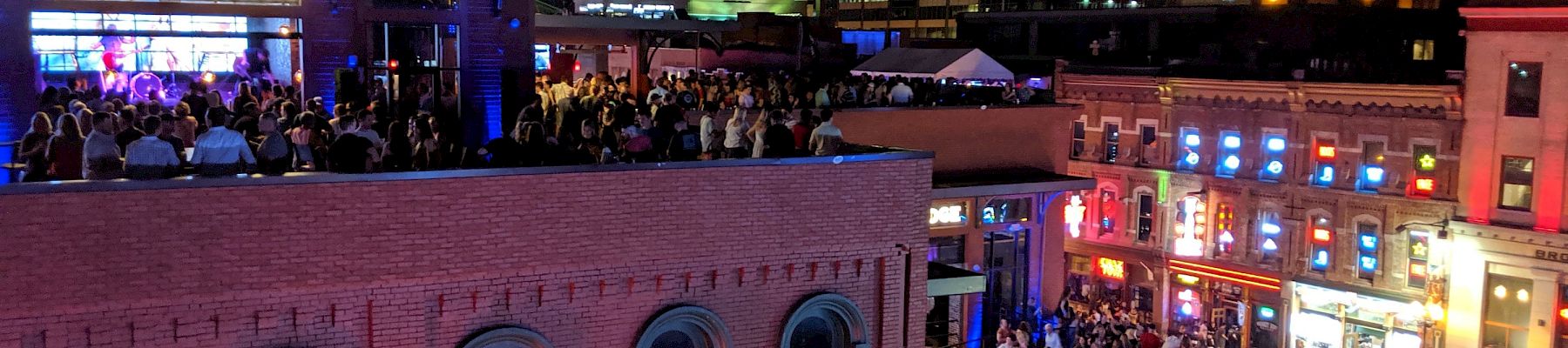 A vibrant nightlife scene with crowds on a rooftop and street, surrounded by brightly lit buildings in an urban area at night.