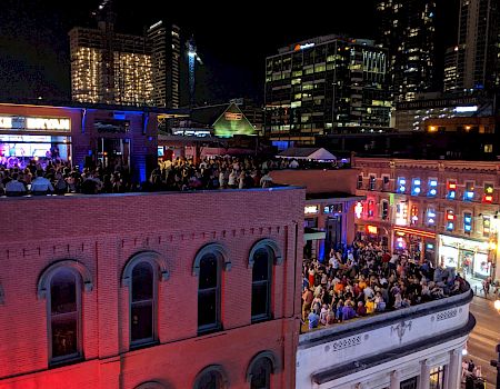 A vibrant nightlife scene with crowds on a rooftop and street, surrounded by brightly lit buildings in an urban area at night.