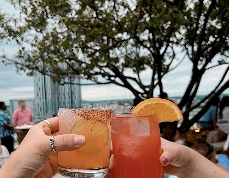 Two hands holding cocktails with citrus garnishes, clinking glasses on a rooftop with a tree and city view in the background.