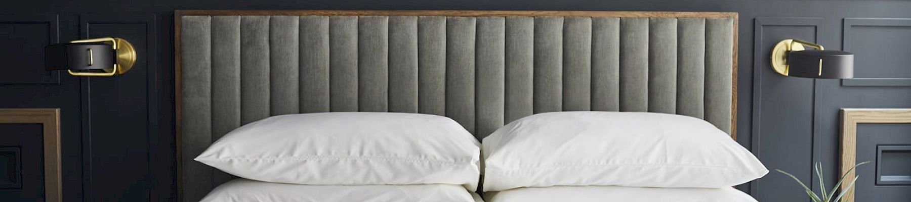 A neatly made bed with stacked pillows, a hat, a blanket, and a newspaper on it, against a dark-paneled wall with brass lights.