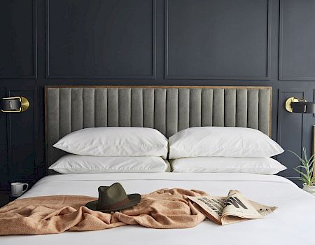 A neatly made bed with stacked pillows, a hat, a blanket, and a newspaper on it, against a dark-paneled wall with brass lights.