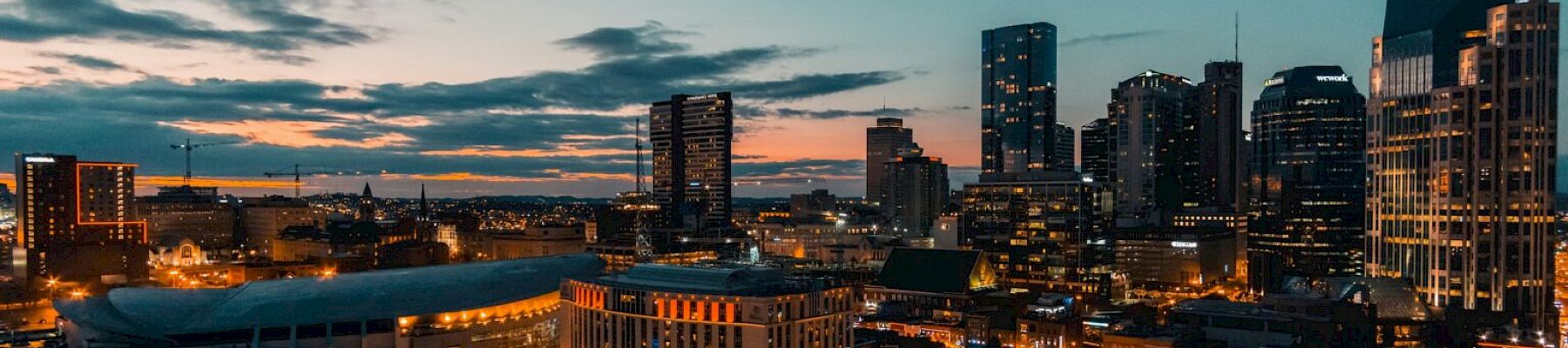 A city skyline at dusk with tall buildings, illuminated streets, and a vibrant sky, creating a vivid urban landscape scene.