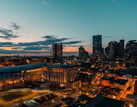 A city skyline at dusk with tall buildings, illuminated streets, and a vibrant sky, creating a vivid urban landscape scene.