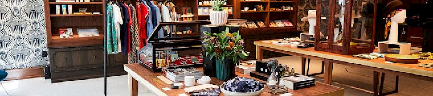 A well-organized boutique with shelves displaying clothes, books, and home decor, featuring wooden tables and a patterned wall.