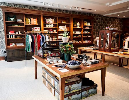 A well-organized boutique with shelves displaying clothes, books, and home decor, featuring wooden tables and a patterned wall.