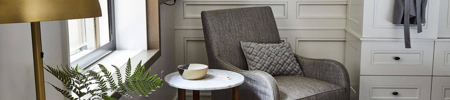 A cozy room features a gray armchair with a pillow, a table with a cup, notepad, lamp, and a robe hanging beside a cabinet.